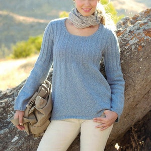 Puede incluir: Un suéter de punto azul claro con cuello redondo y mangas largas. El suéter lo lleva una persona con una bufanda a rayas alrededor del cuello. La persona está de pie frente a una roca grande.