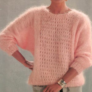 May include: A light pink, open-weave sweater with a round neckline and long sleeves. The sweater has a delicate, airy appearance, with a textured pattern across the front. The sleeves are slightly oversized, and the cuffs are ribbed. A silver bracelet is visible on the wrist.