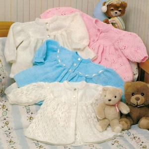 May include: Four knitted baby sweaters in white, pink, blue, and white with a lacy pattern. The sweaters are laid out on a patterned fabric with two teddy bears.