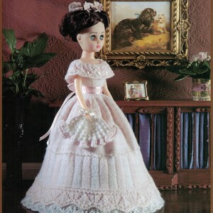 May include: A doll wearing a pink knitted gown with a lace trim. The doll has brown hair and blue eyes. The doll is standing in front of a wooden cabinet with a purple velvet interior.