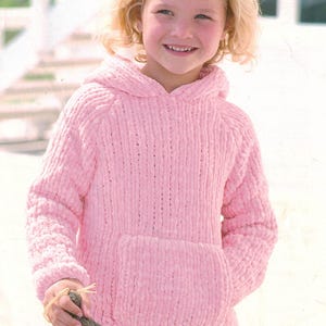 Può includere: Una felpa con cappuccio rosa chiaro con tasca a marsupio. La felpa è realizzata in un tessuto a maglia spesso e strutturato. Il bambino tiene in mano un piccolo bastoncino.