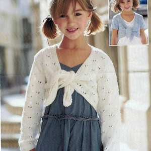 Puede incluir: Una niña lleva un cárdigan blanco de punto, de manga larga y abierto por delante, con un lazo. Lleva un vestido azul. Una imagen más pequeña muestra un cárdigan azul similar.