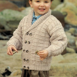 May include: A young boy wearing a light brown knitted cardigan with a wide collar and button fastening. The cardigan has a textured knit pattern and is worn over a blue shirt. The boy is holding a small object in his hand.