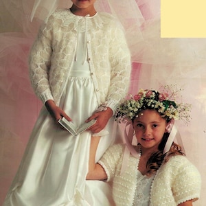 May include: Two young girls in white dresses and matching lace-trimmed sweaters. One girl wears a floral crown and the other a veil. Both are dressed for a special occasion, possibly a wedding or a religious ceremony.