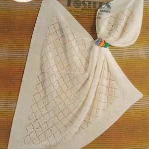 May include: Cream-colored knitted baby blanket with a diamond pattern and a decorative border. The blanket is folded and held together with a colorful ring. The background is a striped pattern in shades of brown and yellow.