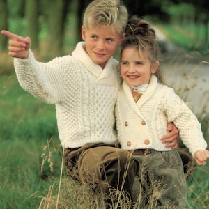 May include: Two children wearing white knitted cardigans with a collared neckline. The boy is pointing to the right and the girl is smiling. They are standing in a grassy field.