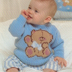 Puede incluir: Un bebé con un suéter de punto azul con un diseño de oso de peluche marrón. El suéter tiene un lazo blanco en el oso de peluche. El bebé está sentado en una manta blanca.