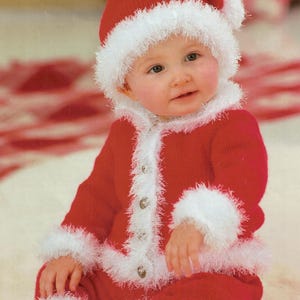 May include: A baby wearing a red Santa Claus outfit, including a hat, jacket, and pants. The outfit is trimmed with white faux fur. The baby is sitting and smiling.