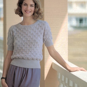 May include: A woman wearing a light grey knitted sweater with a delicate openwork pattern and a matching grey skirt.