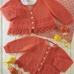 May include: Two coral-colored knitted baby cardigans with white collars and button closures. The cardigans feature a decorative scalloped hem and a detailed knit pattern. The image includes decorative fruit and a red and white checkered background.