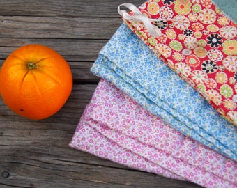 May include: Three fabric tea towels with floral patterns. The top towel is red with white and yellow flowers. The middle towel is blue with white flowers. The bottom towel is pink with white flowers. An orange sits on a wooden surface.