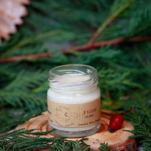 Puede incluir: Un pequeño frasco de vidrio con una crema blanca en su interior. El frasco tiene una etiqueta marrón con el texto "A VISTA (OLIRES) Chamomilla Nerva Nostrum". El frasco está sobre una superficie de madera con follaje verde en el fondo.
