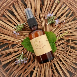 Op de afbeelding: Een bruine glazen fles met een spuitmond en een bruin papieren etiket. Het etiket heeft een halve maan en de tekst "Artemisia - Alchemy". De fles staat op een geweven rieten oppervlak.