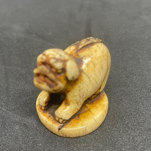 May include: A small, intricately carved ivory figurine of a dog, with a detailed face and a curled tail. The figurine is sitting on a round base.
