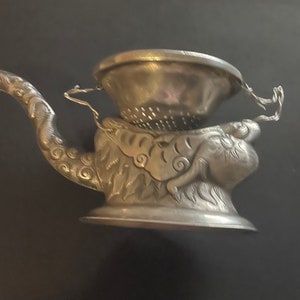 Può includere: Teiera d'argento con un designo a forma di drago e un coperchio con filtro. La teiera ha un beccuccio e un manico.