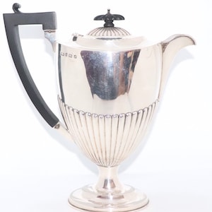 Puede incluir: Una cafetera de plata con asa negra y diseño acanalado. La cafetera tiene un cuerpo redondo y un pico alto y delgado. La tapa tiene un pomo negro.