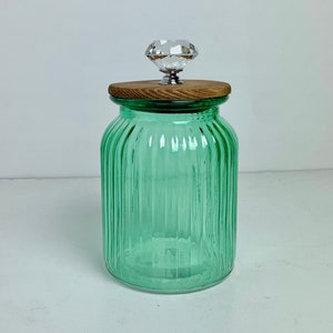 May include: A green glass jar with vertical ribbed detailing. The jar has a wooden lid and a clear, faceted crystal knob. The jar is empty and sits on a white surface.