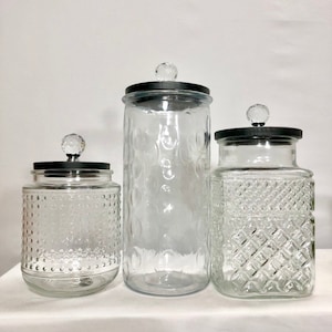 May include: Three clear glass jars with decorative lids. The jars vary in size and have different textured patterns, including a dotted, circular, and diamond design. Each jar has a black lid with a crystal-like knob on top. The jars are likely for kitchen or home decor.