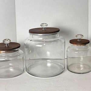 May include: Three clear glass jars of varying sizes with wooden lids and crystal-like knobs. The jars are empty, showcasing their transparency. The lids are a dark brown color, and the knobs are clear and faceted.