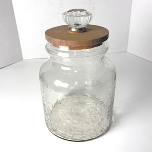 Könnte beinhalten: Ein klares Glasgefäß mit strukturierter Oberfläche und einem Holzdeckel. Der Deckel ist mit einem transparenten, dekorativen Knopf versehen. Das Gefäß ist leer und steht auf einer weißen Oberfläche.