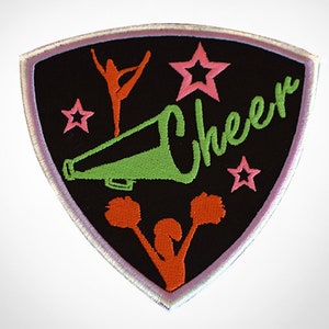 May include: Embroidered patch with a black shield shape, featuring the word "Cheer" in green script. Includes orange cheerleader figures, pink stars, and a green megaphone. The patch has a light purple border.