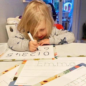 May include: A child in a grey sweatshirt with star motifs is writing numbers with a black marker on a white sheet of paper. The numbers 0 to 7 are printed on the paper. Other sheets of paper with the name "Emma" are also visible.