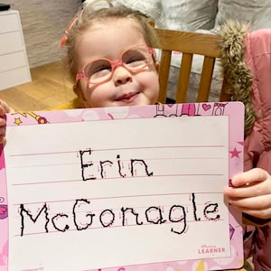 May include: A young girl wearing pink glasses holds up a pink and white dry-erase board with the words "Erin McGonagle" written in black marker on it. The board has pink lines and a pink castle design at the top.