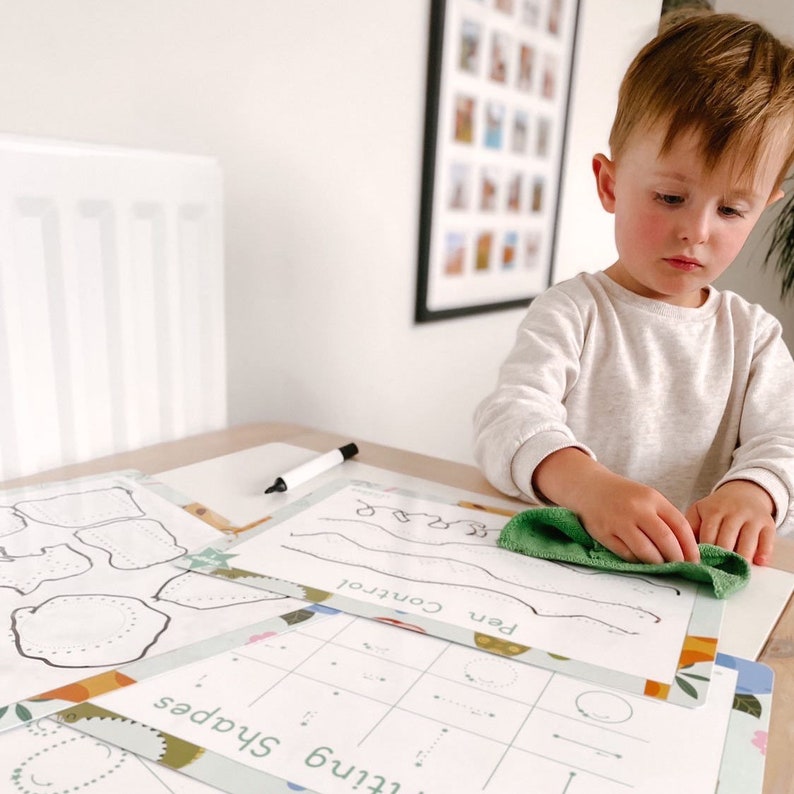 May include: A child is working on a set of printable worksheets with a green wipe. The worksheets include activities for learning shapes, pen control, and fine motor skills. The worksheets are printed on white paper with a green and orange border.
