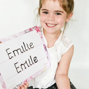 May include: A young girl holds up a pink and white dry-erase board with the name "Emilie" written on it twice. The board has a unicorn theme with pink stars and hearts.