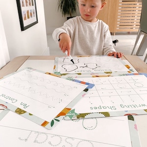 May include: A child is using a white marker to trace shapes on a white dry-erase mat. The mat has a green and orange border with a floral pattern. The mat says "Tracing Shapes" and "Stage 1".