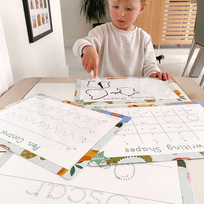 May include: A child is using a white marker to trace shapes on a white dry-erase mat. The mat has a green and orange border with a floral pattern. The mat says "Tracing Shapes" and "Stage 1".