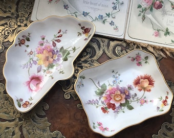 May include: Two white porcelain decorative dishes with gold trim. The dishes feature floral patterns in shades of pink, yellow, and purple. The larger dish is shaped like a shell and the smaller dish is rectangular.
