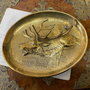 May include: An oval brass tray featuring a raised, three-dimensional relief of a stag head in the centre. The surface of the tray has a textured, stippled finish surrounding the deer, while the edges are smooth and polished with a metallic gold tone.