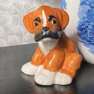 Può includere: Statuetta di cane boxer in ceramica, seduta. Il cane ha un mantello marrone e bianco con dettagli neri sul naso e sugli occhi. La statuetta è su una superficie nera.