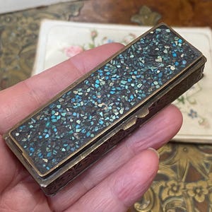 May include: A rectangular antique box with a mosaic top. The mosaic features small, irregular pieces in shades of blue, turquoise, and white, set against a dark background. The box has a brass frame and decorative etched sides.