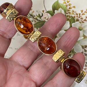 May include: A gold-tone link bracelet featuring four large, oval-shaped, translucent amber-colored cabochons. Each amber stone is set in a smooth gold-tone bezel, alternating with rectangular gold-tone links that display an engraved swirl pattern.