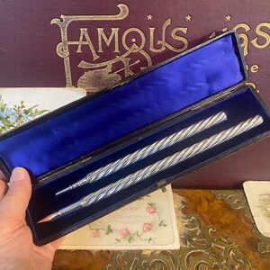 May include: Silver pens with a twisted design are presented in a black case with a blue velvet interior. The pens are in a rectangular case with a hinged lid. The case is held open by a hand.