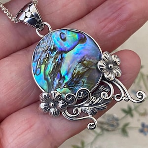 May include: Abalone shell pendant necklace with a silver chain. The round pendant features iridescent blues, greens, and purples. The pendant is accented with silver floral and leaf designs. The necklace is held in a hand.