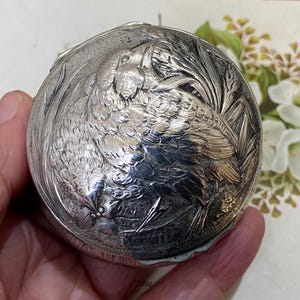 May include: A round silver-toned metal container featuring a detailed embossed design of a bird perched among foliage on its lid. The surface has a polished, reflective finish with intricate textures depicting feathers and plant leaves.