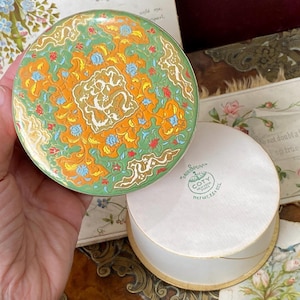May include: A vintage Coty powder compact with a vibrant, ornate design in green, orange, and white. The compact is round and has a matching base. The text "Coty" is visible on the base.