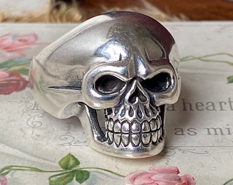 Könnte beinhalten: Ein silberner Totenkopfring mit einem detaillierten Design. Der Schädel hat eine ausgeprägte Stirn, tiefe Augenhöhlen und ein zahniges Grinsen.