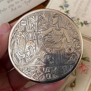 May include: A silver-coloured, round compact with an engraved Egyptian design. The design features a seated figure, a cobra, and floral elements. The compact is held in a hand, with a blurred background of paper and flowers.