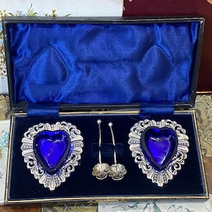 May include: An antique set of serving pieces in a presentation box. The set includes two heart-shaped dishes with cobalt blue glass inserts and ornate silver-toned frames, plus two small serving spoons. The box has a blue velvet lining.