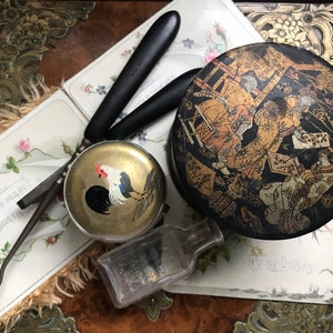 May include: A vintage round tin box with a black background and gold painted Asian figures. The box is on a wooden surface with a small bottle, a hair pick, and a round tin box with a rooster design.