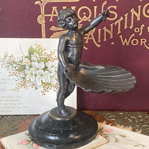 Puede incluir: Una escultura en bronce de una figura de querubín sosteniendo una gran concha, montada sobre una base de mármol negro. La figura apunta hacia arriba. El fondo incluye postales antiguas y un libro titulado "Famous Paintings of the World."