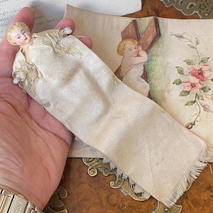 May include: A small antique doll with a porcelain head and painted features, dressed in a cream satin gown. The doll is held over a vintage card with a painted cherub and floral design. The card has a cream satin ribbon attached.