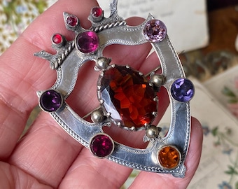May include: A silver-toned brooch with a large, faceted, reddish-brown central stone. The brooch is adorned with various colored gemstones, including red, pink, purple, and orange. The design is ornate and features decorative elements.