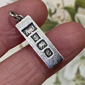 May include: A rectangular silver pendant featuring a textured surface and four distinct stamped hallmarks arranged vertically. The stamps include the letters AJ, an anchor symbol, a lion passant figure, and the letter L, all set against a dark background.