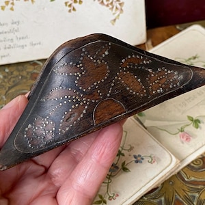 May include: A dark brown wooden sewing tool with a triangular shape, featuring an inlaid floral pattern created with small metal pins. The wood has a smooth, polished finish and the metal pin design depicts a central flower with leaves.