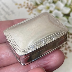 Puede incluir: Una pequeña caja rectangular de plata con una tapa pulida y lisa que presenta un borde decorativo en relieve. Los lados del recipiente son lisos y reflectantes, mostrando un acabado metálico con detalles intrincados.